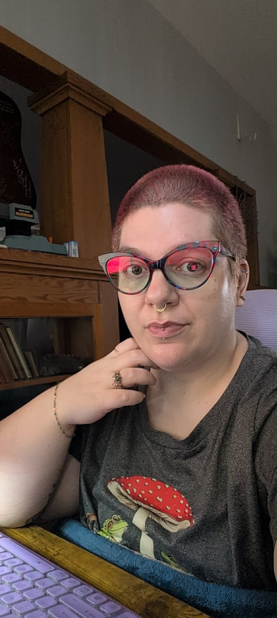 Ruth Kearney sitting at a desk with a purple keyboard, wearing cat-eye glasses, a septum piercing, and a grey t-shirt featuring a frog next to a red-and-white mushroom.